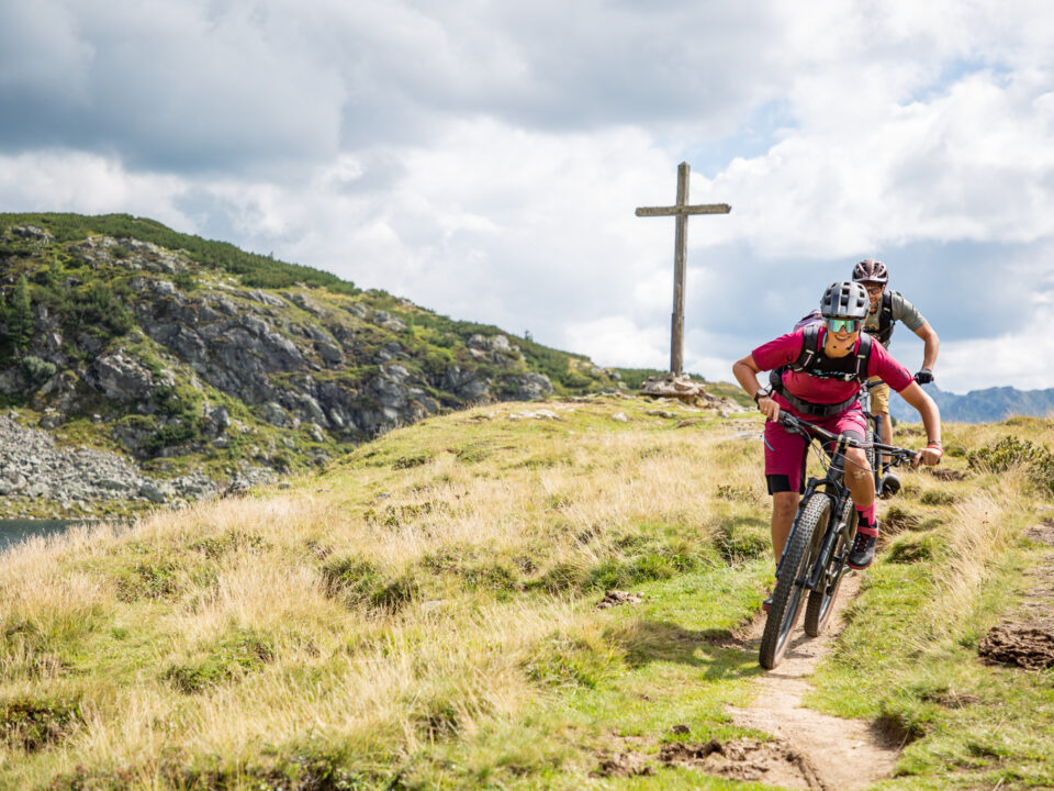 2 Mountainbiker, die im Bereich der Oberhütte in Obertauern auf dem Trail mit dem Mountainbike unterwegs sind. Im Hintergrund ein Teil des Oberhüttensees, die Berge und ein Gipfelkreuz.