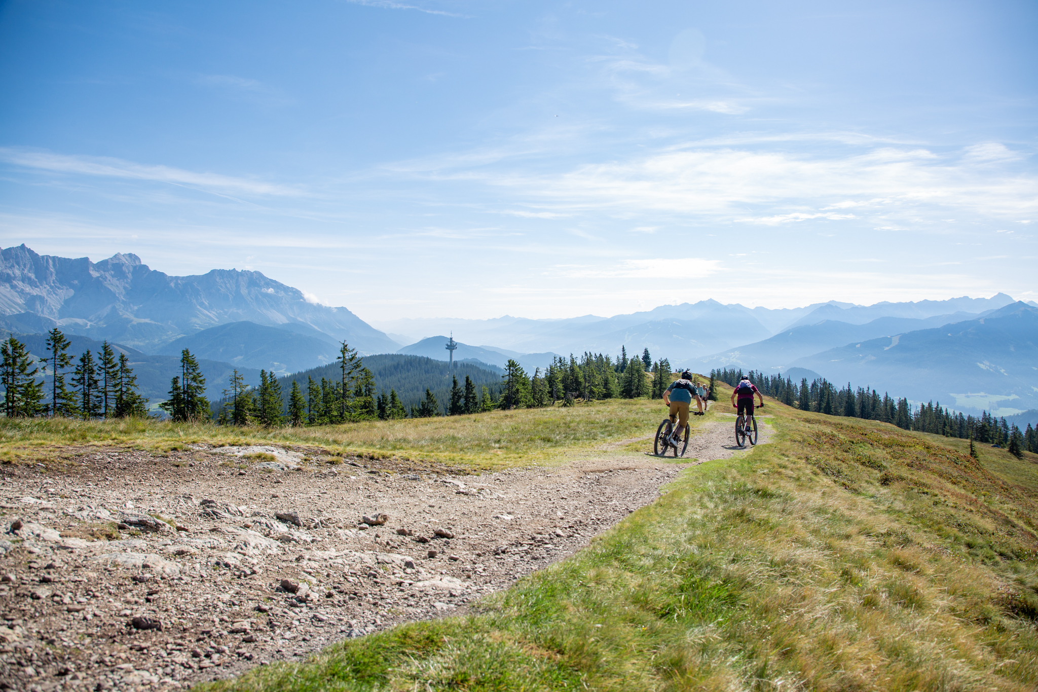 Die ultimative MTB-Challenge Stoneman Taurista. Auf dem Bild sieht man zwei Mountainbiker von hinten in der Nähe des Rossbrandgipfelkreuzes, im Hintergrund zahlreiche Berge in und rund um die Salzburger Sportwelt.
